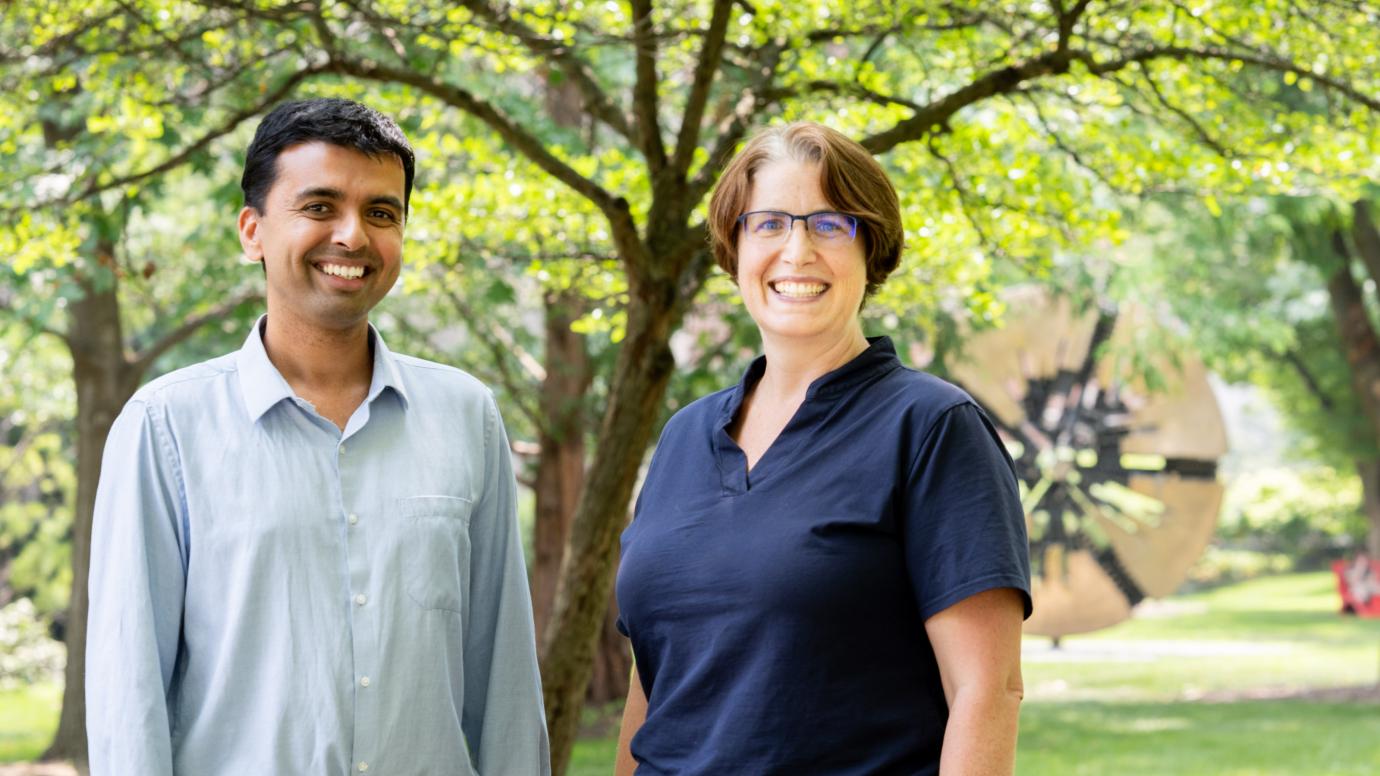 Two people smile and stand by a tree